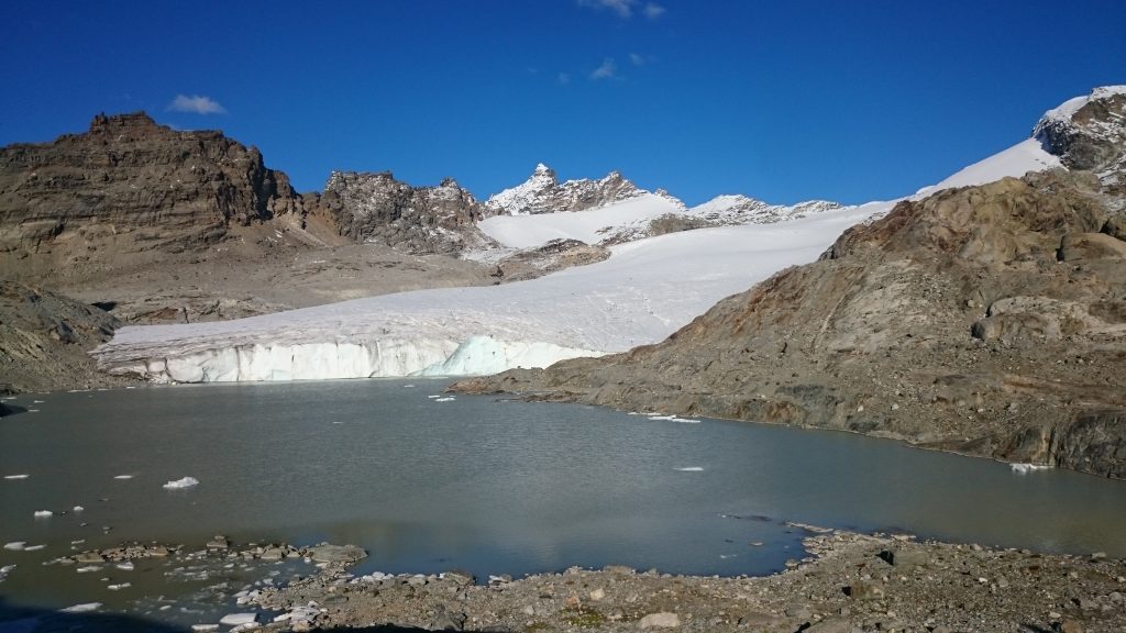 Au premier plan un lac glaciaire, issu de la fonte du glacier du Grand méan, situé juste derrière. Au fond des sommets alpins de 3000m qui délimitent la frontière avec l'Italie.