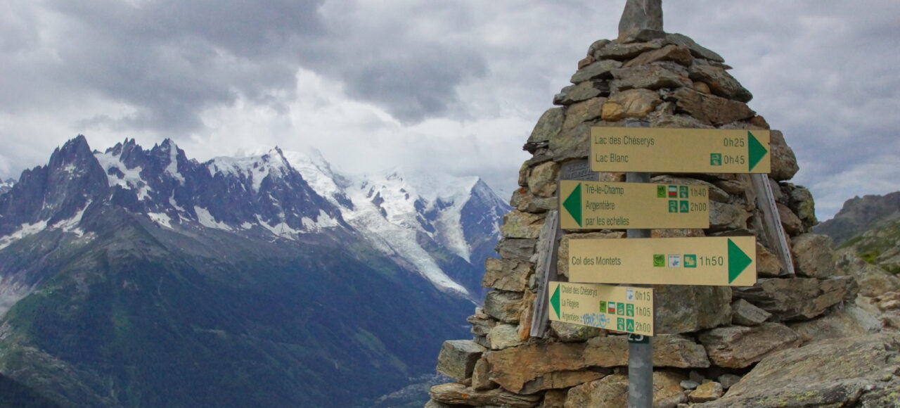 panneau indicatif situé devant un cairn massif, avec en arrière plan des montagnes et le mont-blanc